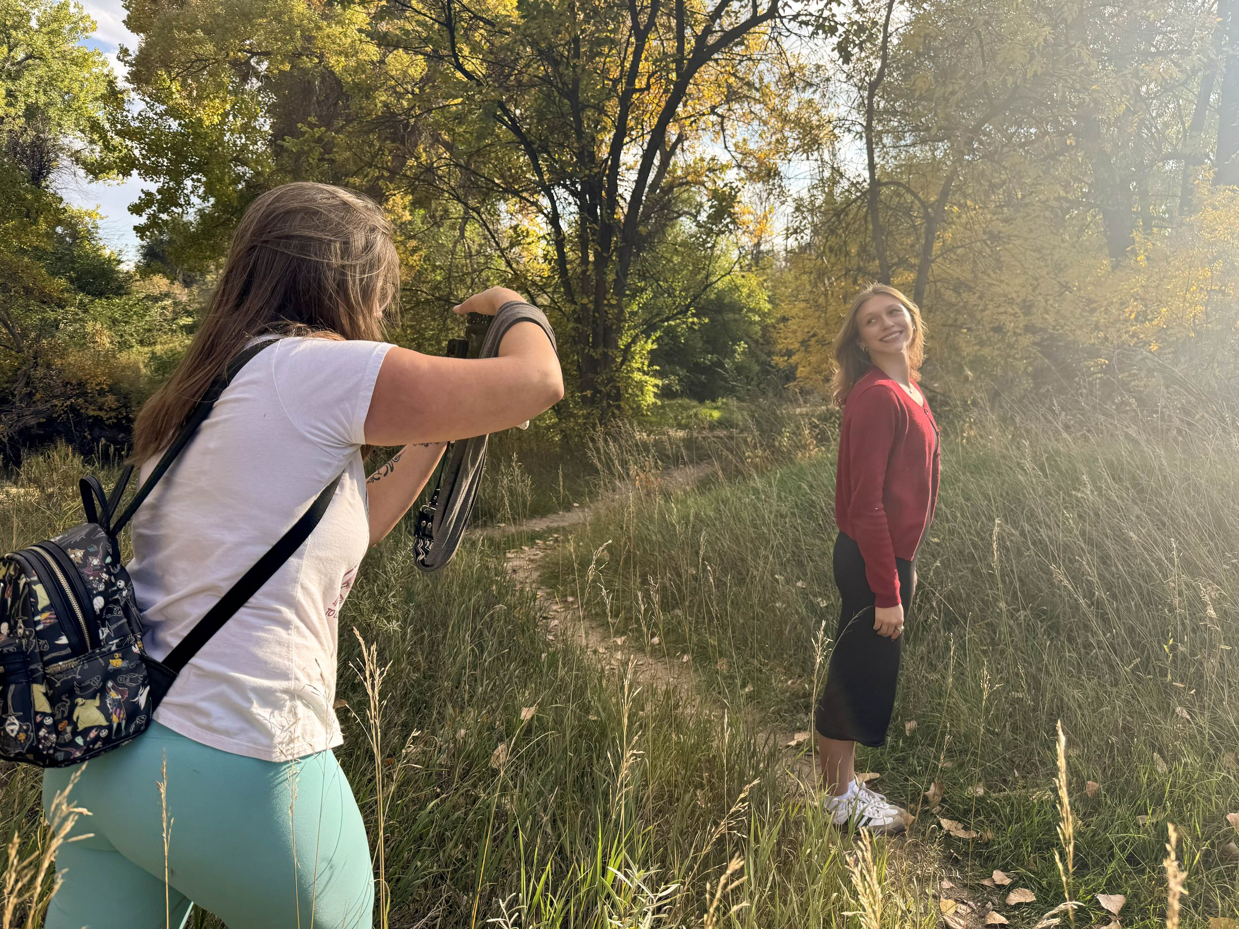 A young woman photographs a girl posing on a nature trail surrounded by tall grass and autumn trees in golden sunlight.