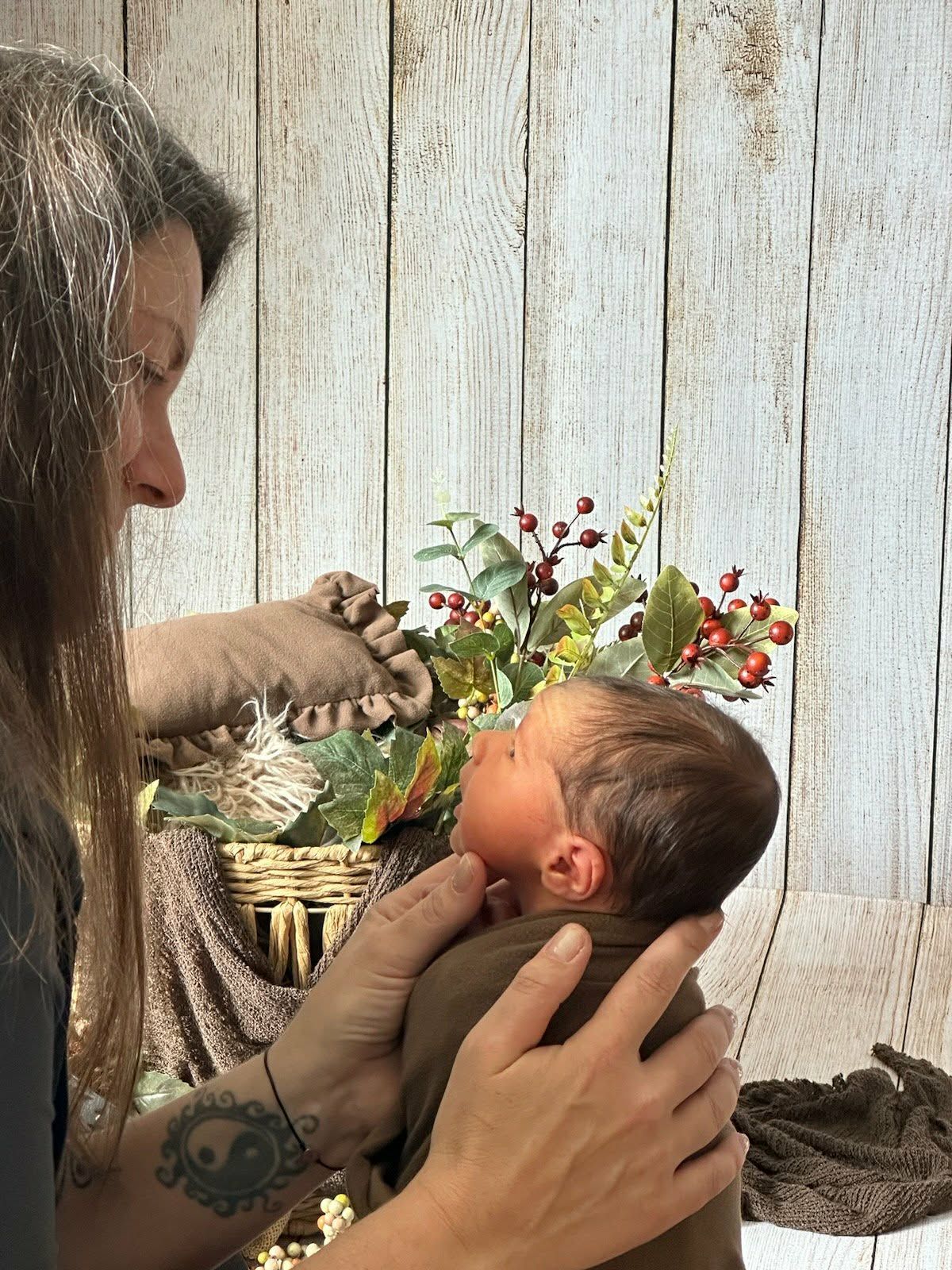 A woman with tattoos tenderly holds a newborn baby near a decorative basket with autumn berries and foliage.