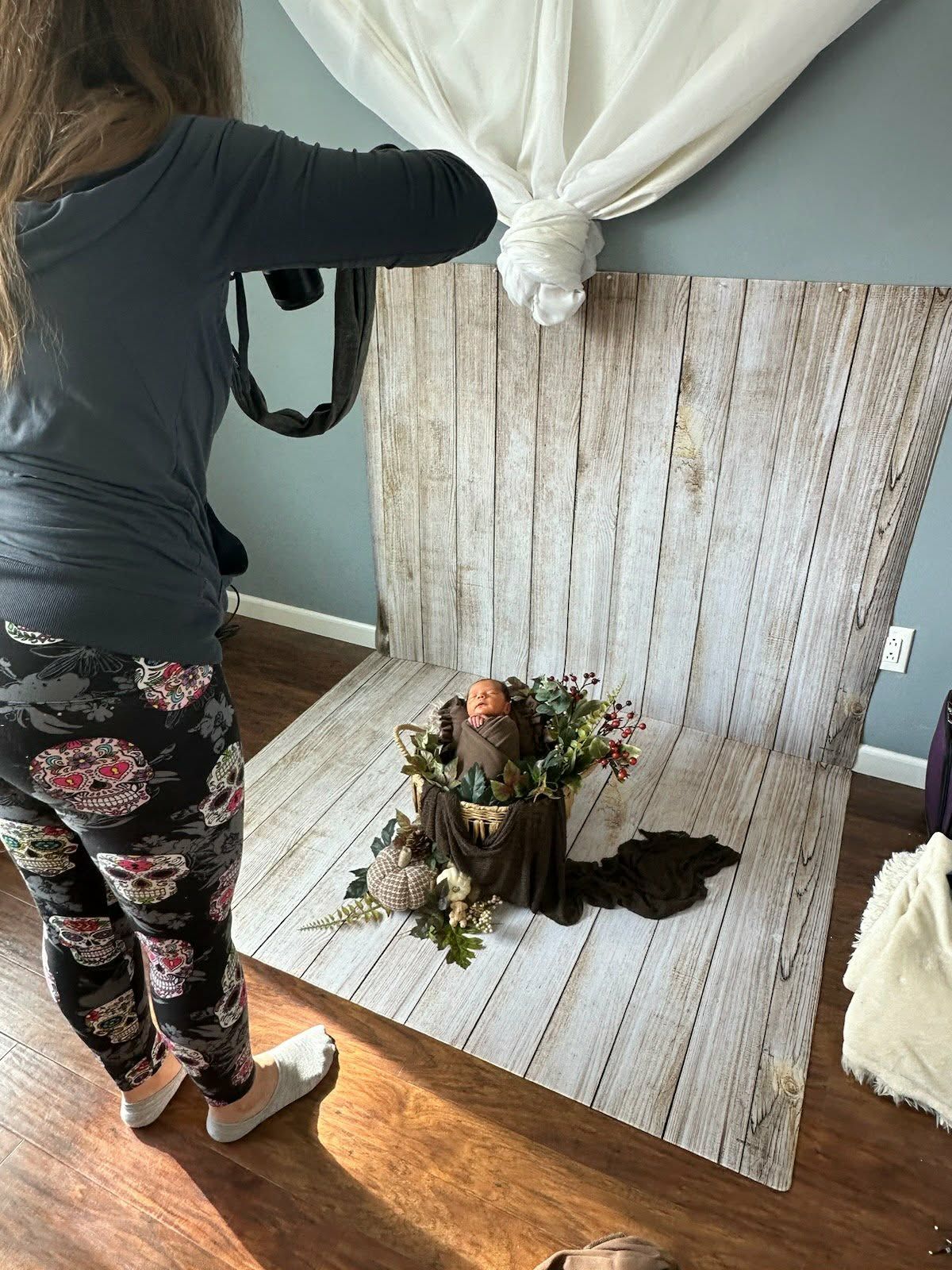 Photographer capturing a newborn baby posed in a floral basket setup against a rustic white wood backdrop in a studio.