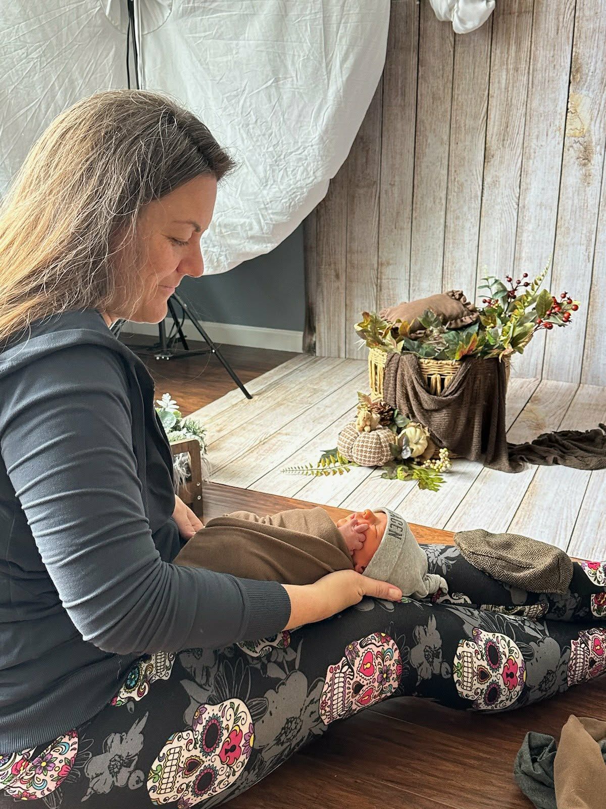Woman in skull-print leggings sits on floor holding a newborn baby in a photography studio with fall floral decor.