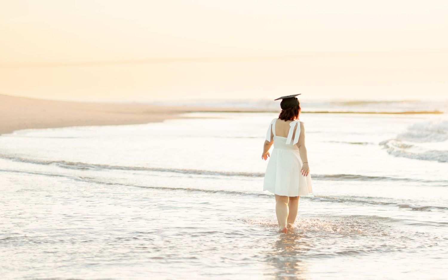 Two hands hold a graduation cap with gold tassel and white dress on beach