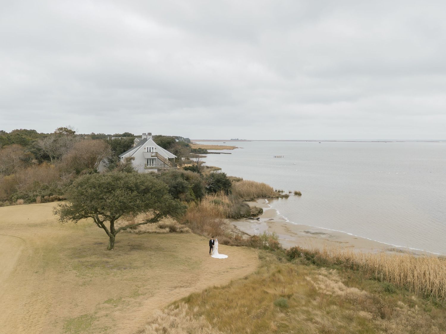 Aerial view of bride and groom on coastal shoreline near rustic waterfront cottage.