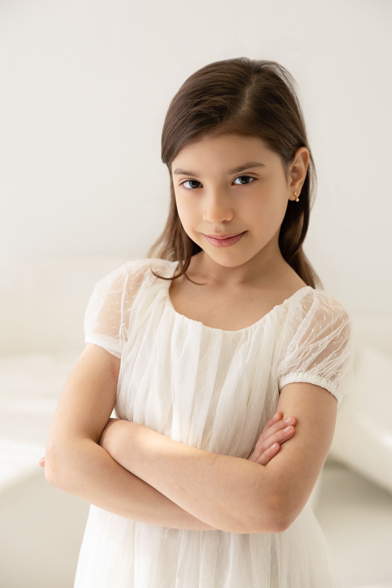 Young girl in white lace dress poses with crossed arms against light background.