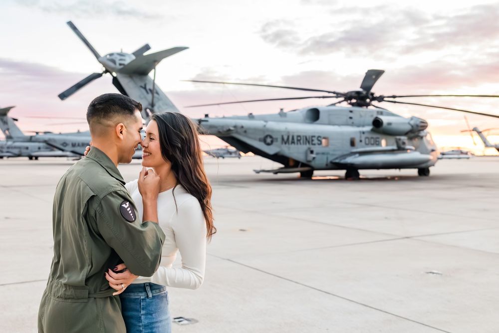 Military couple embraces on airfield with helicopters in background during sunset.