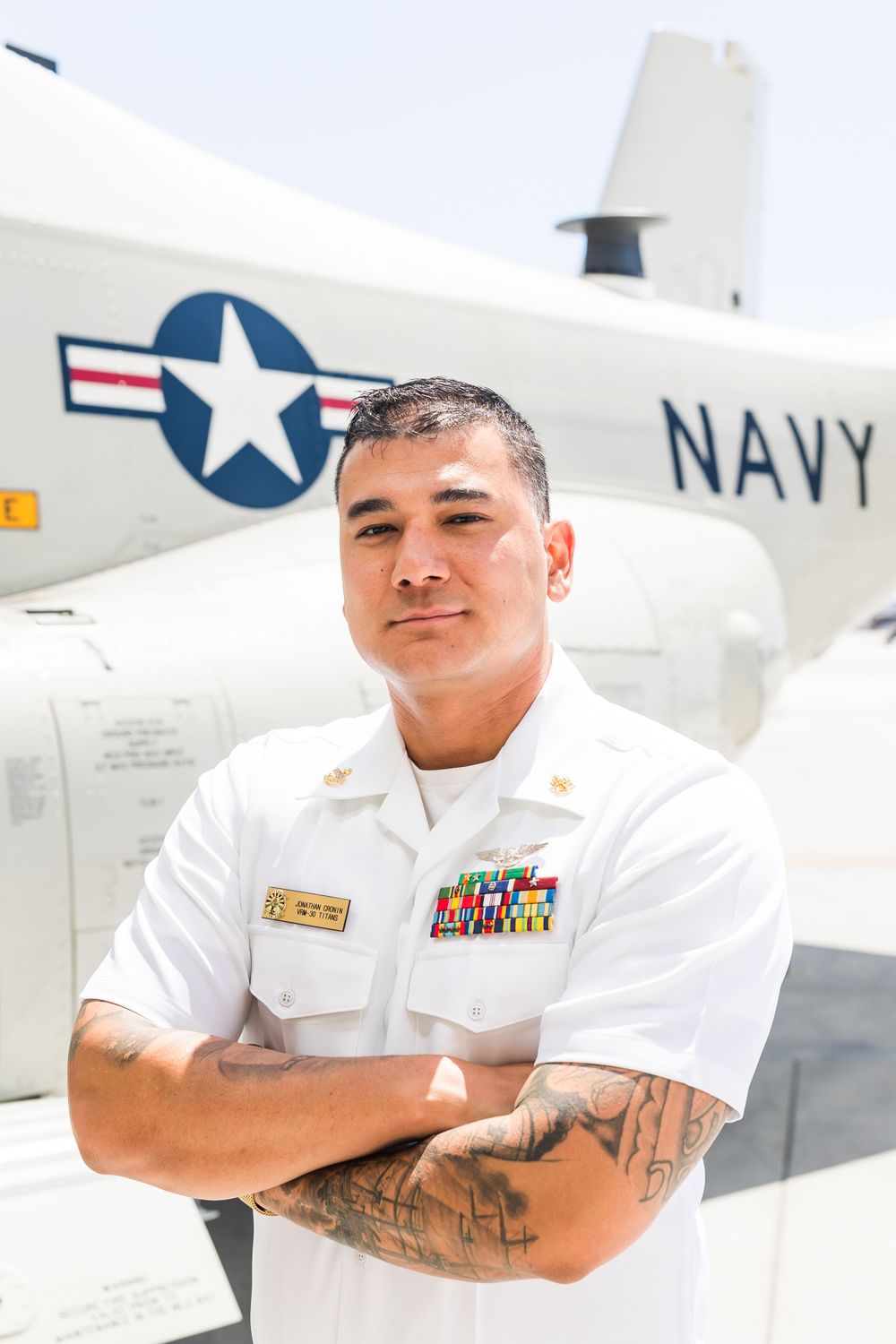 A Navy service member in white uniform standing with crossed arms in front of a military aircraft with Navy markings.