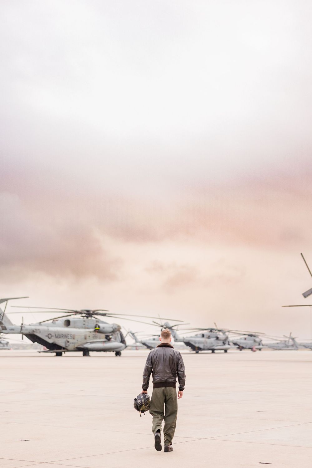 Several military helicopters and planes parked on a misty airfield at sunrise.