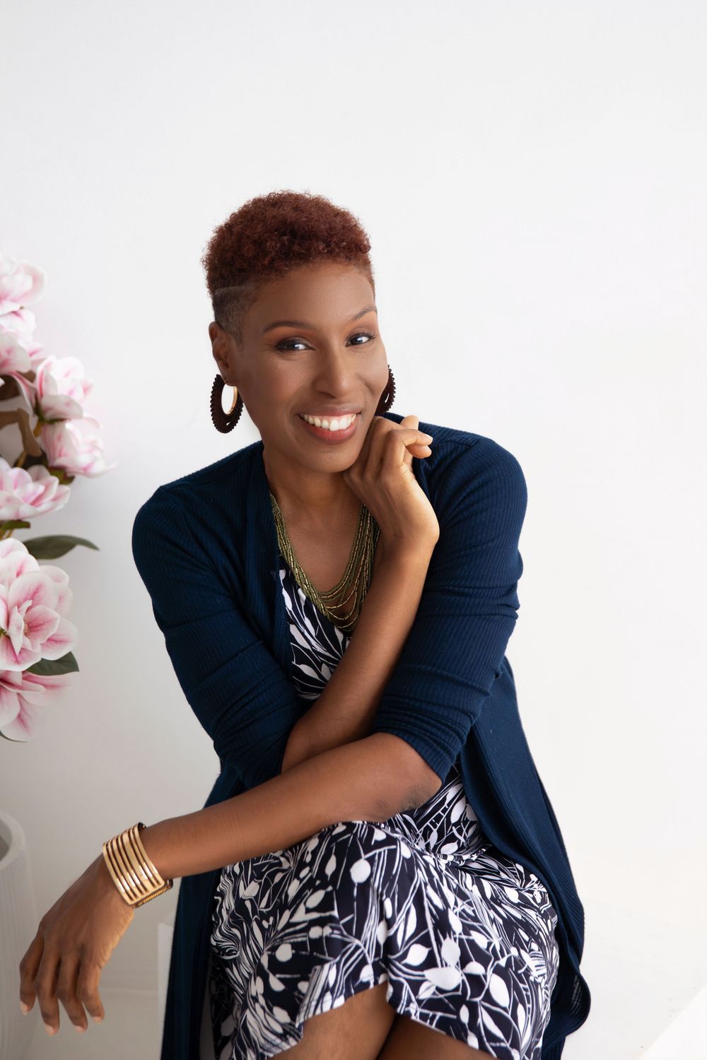Stylish woman in navy cardigan and floral dress poses elegantly against white backdrop with pink flowers.