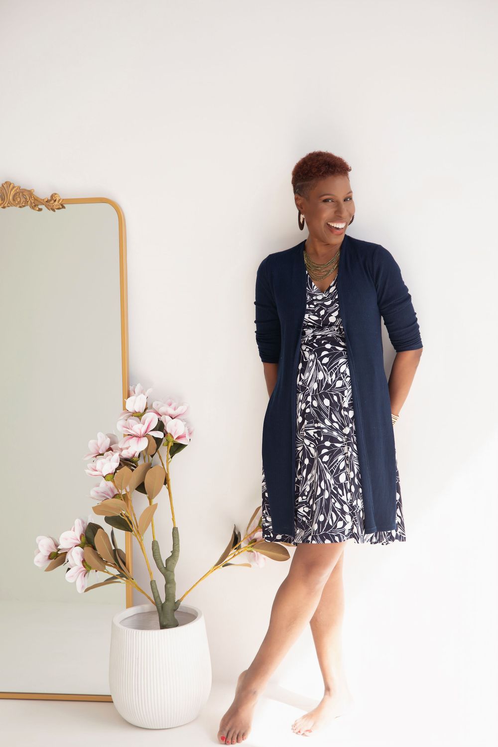 A person wearing a navy cardigan over a floral dress stands barefoot next to a white vase with pink flowers.