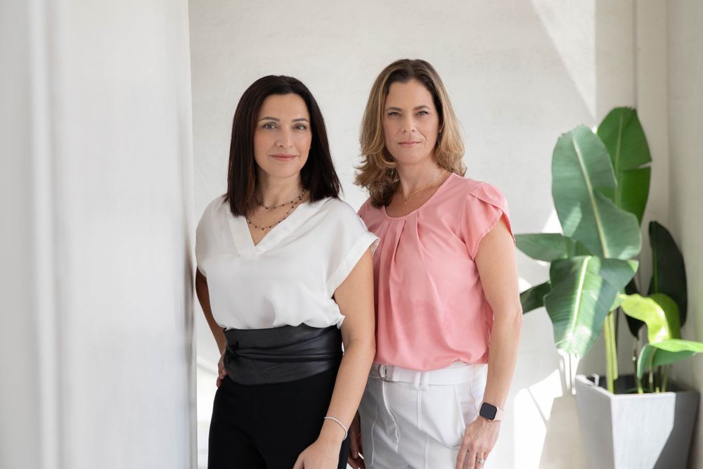 Two women in casual business attire standing against white wall with large indoor plant.