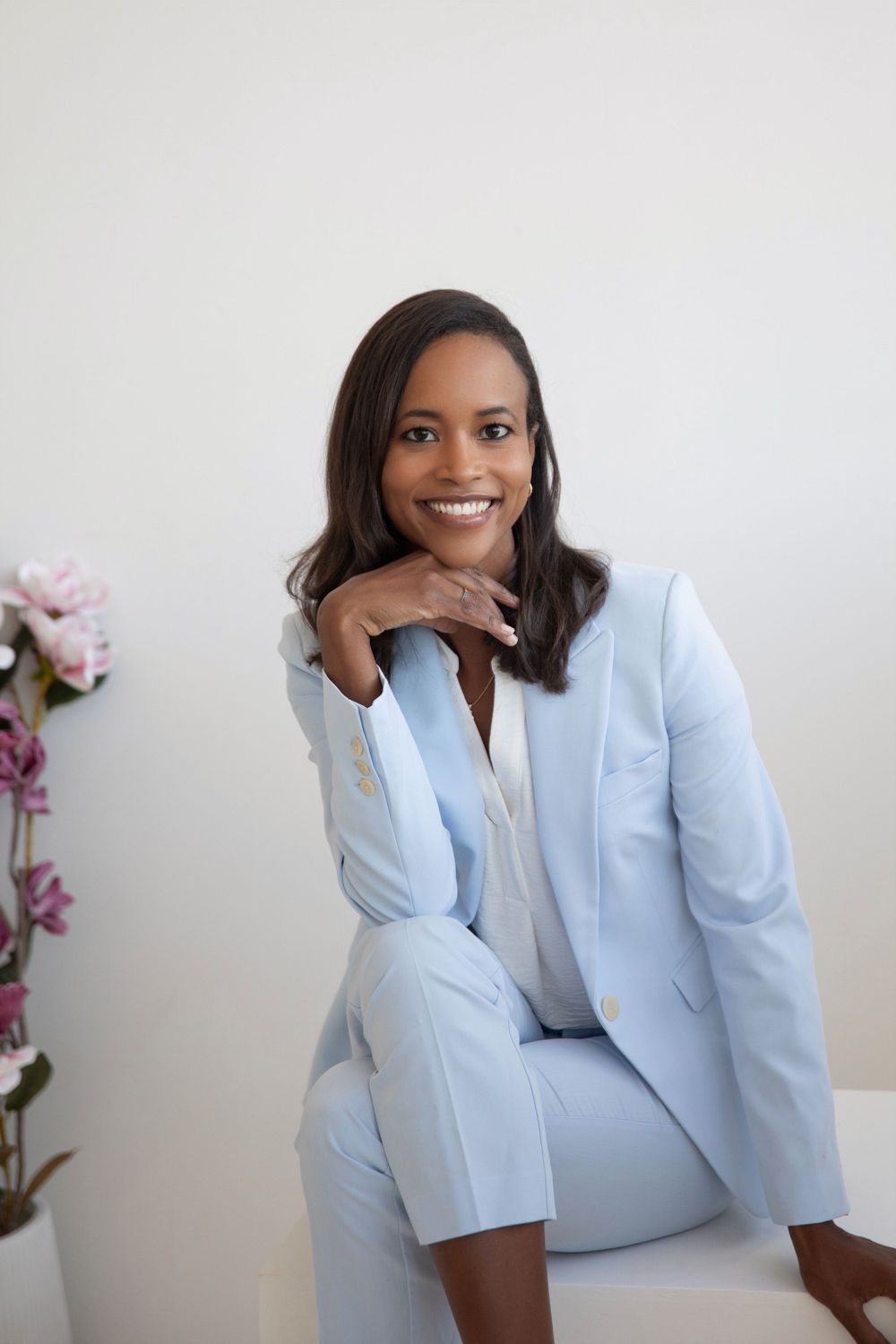 Professional wearing a light blue suit sits confidently on a white bench with pink flowers in the background.