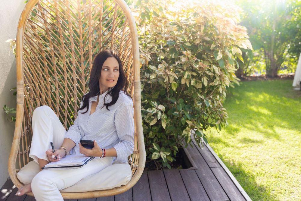Person relaxes in hanging rattan chair on deck with wine glass amid sunlit garden setting.