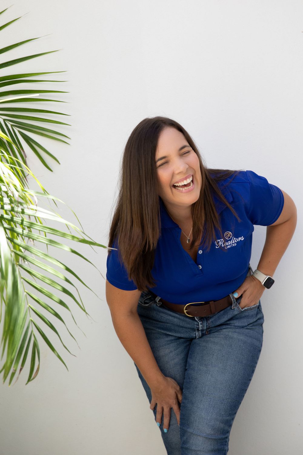 Woman in blue polo shirt and jeans laughing against white wall with palm fronds.