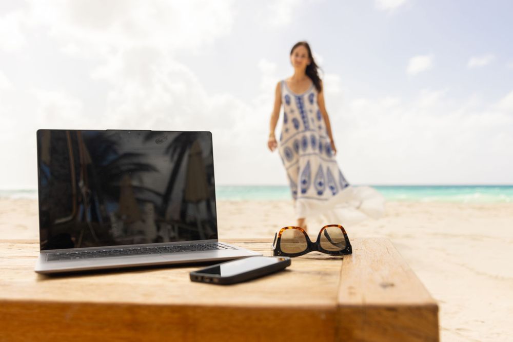 Laptop and sunglasses on wooden table at beach with blurred person and turquoise waters in background.