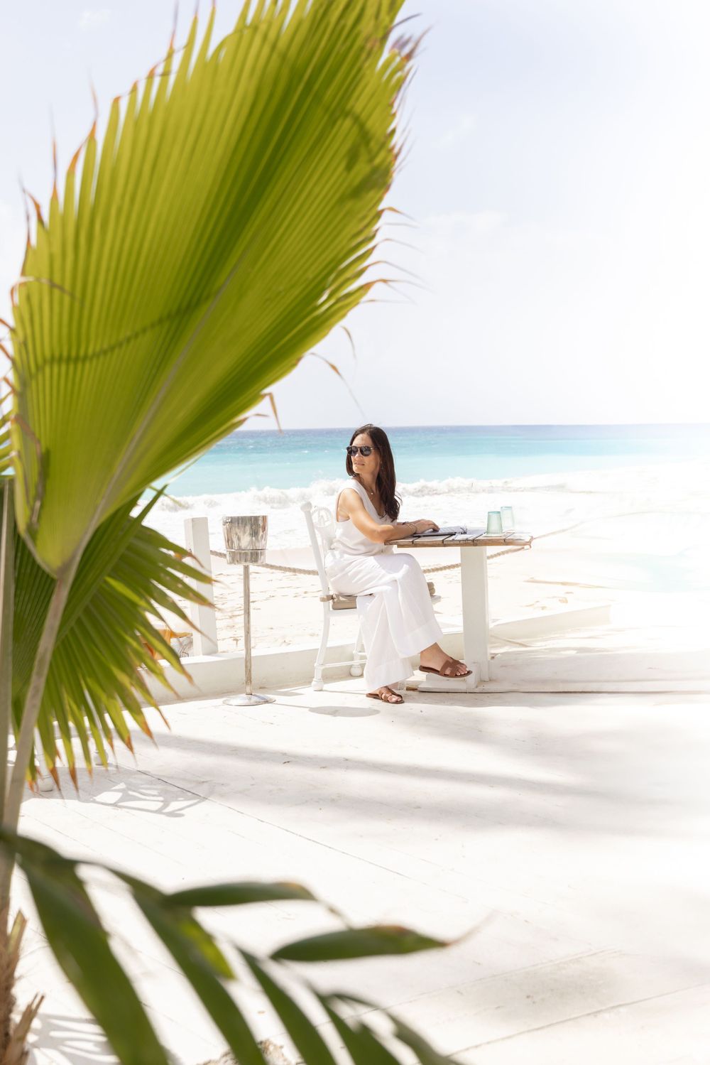 A beachside terrace dining setup with a palm frond in the foreground and turquoise ocean waters in the background.