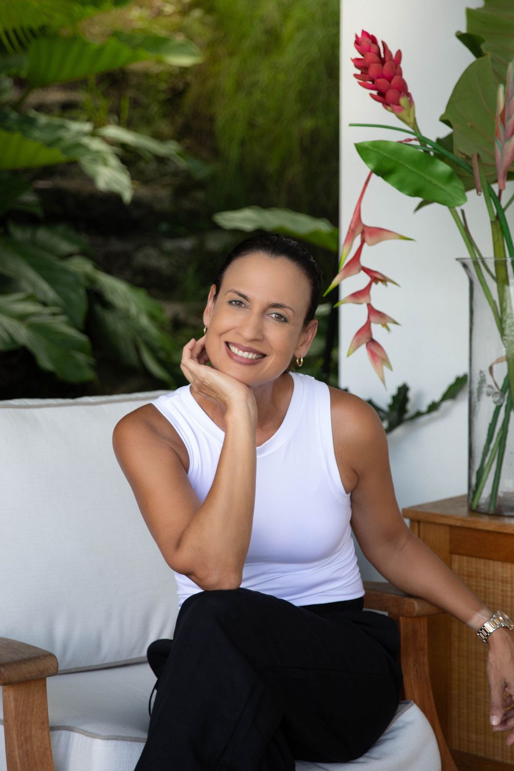 Woman in white tank top relaxing on outdoor patio with tropical flowers and plants.