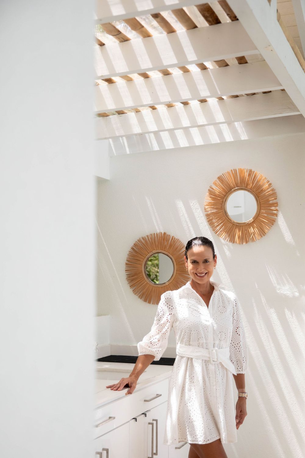 White lace dress interior shot with natural light streaming through wood beams and rattan sunburst mirrors on wall.