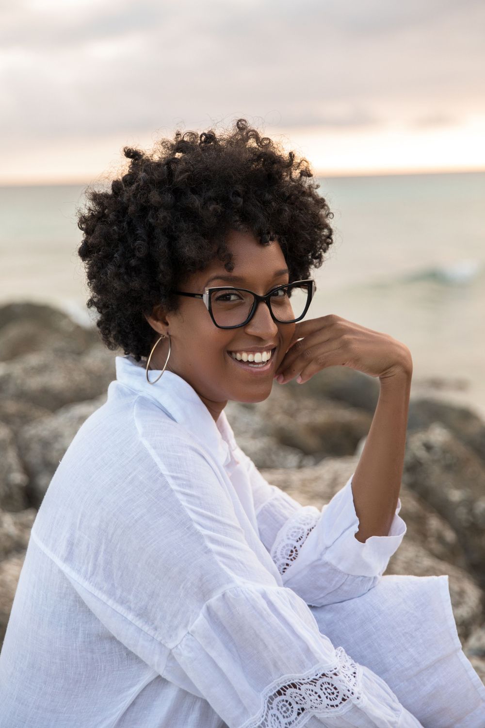 Portrait of person in white lace dress sitting on coastal rocks at sunset.