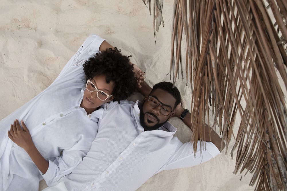 Couple in white clothing relaxing together under palm fronds on sandy beach.