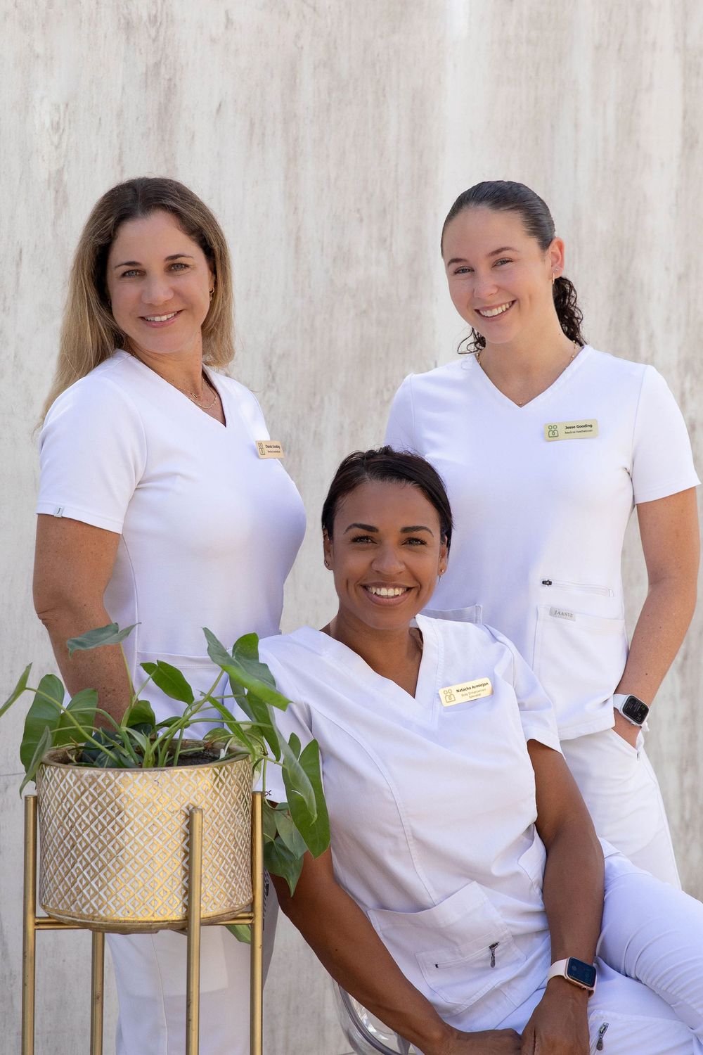 Three healthcare workers in white uniforms posing together with decorative plant.