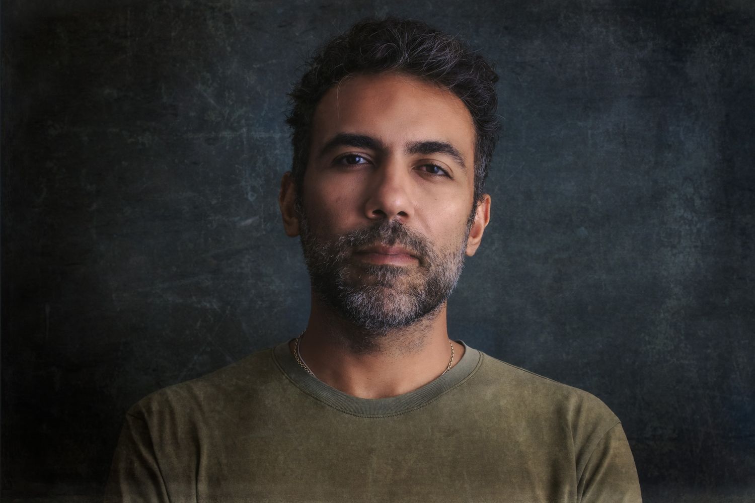 Bearded man in olive green t-shirt posing against dark textured background.