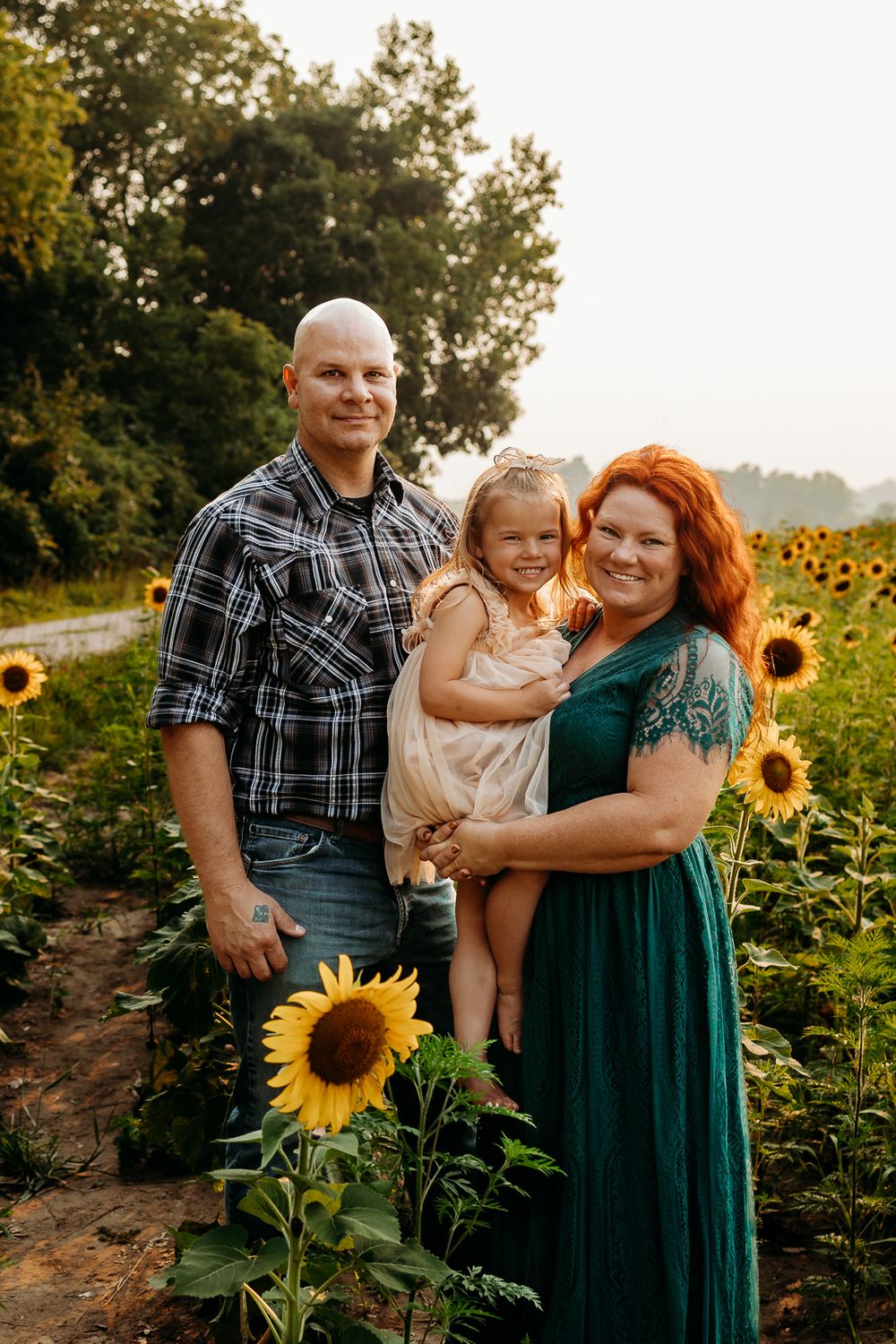 A mom, dad, and little girl all standing together in a field of sunflowers smiling.