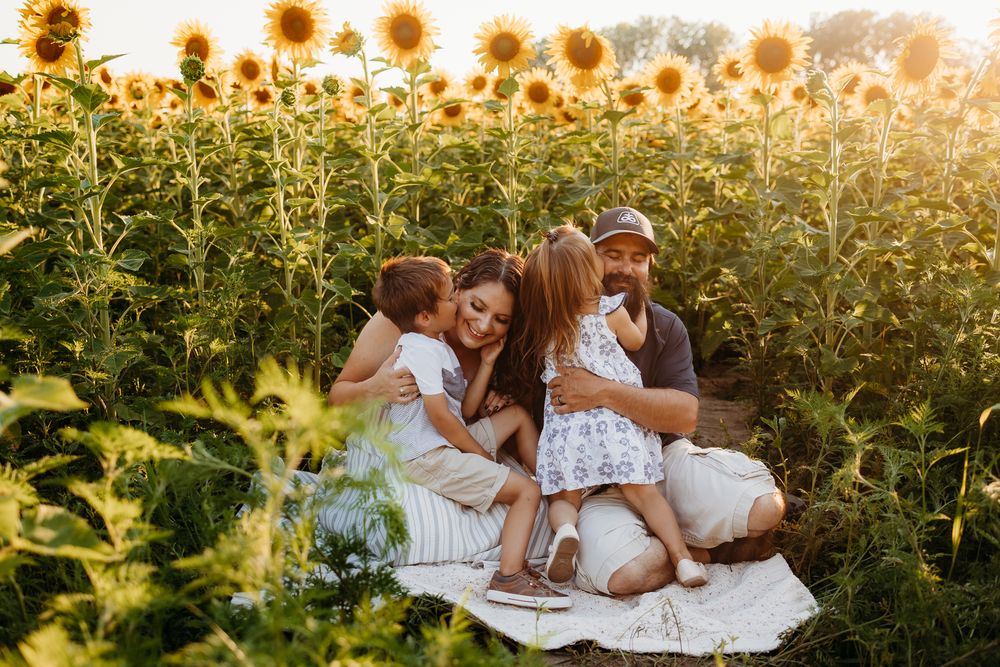 A family of four sitting on a blanket in a field of sunflowers. The kids are giving their parents a kiss on the cheek.