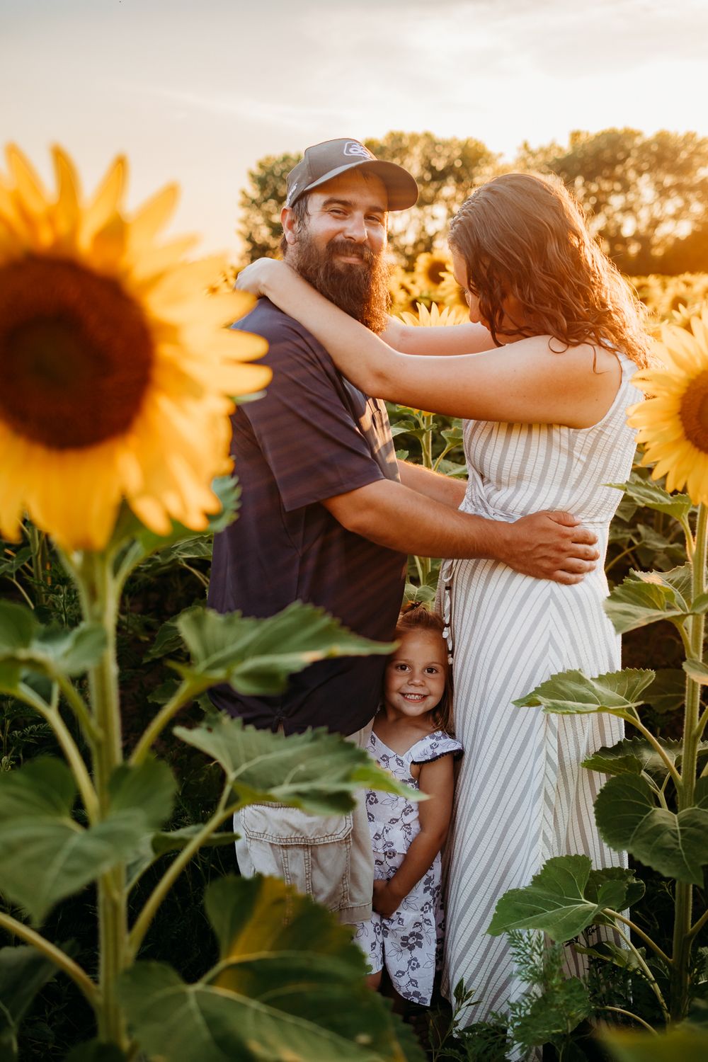 A couple embracing each other in a field of sunflowers as their daughter is standing between them smiling.