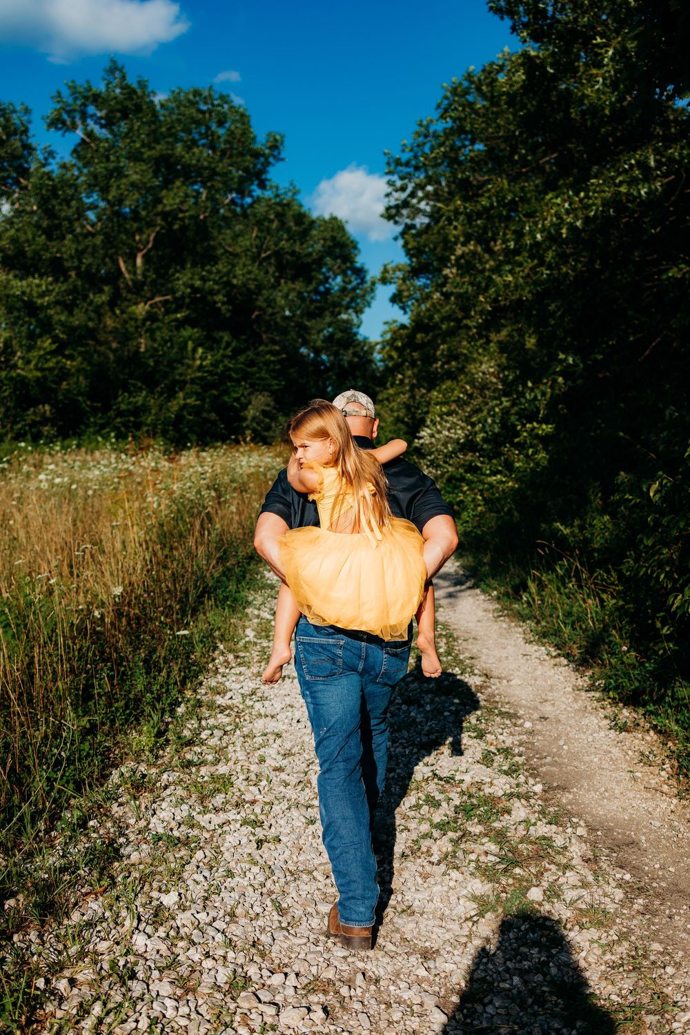 A dad giving his daughter a piggy back ride on a gravel path through a field.