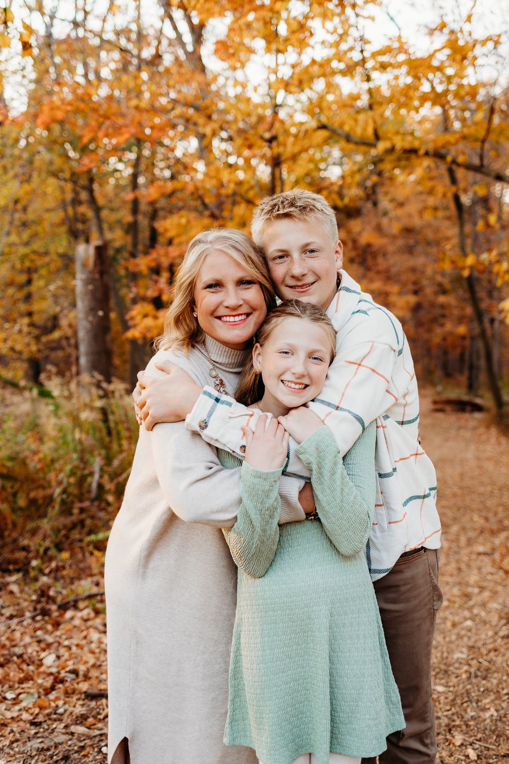 A family of three all hugging each other on a trail in the forest. There are leaves in the ground and the trees have changed colors.