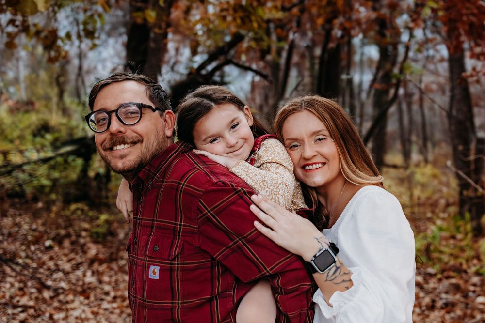 A Mom and Dad and their daughter all smiling for a picture. Leaves are on the ground.