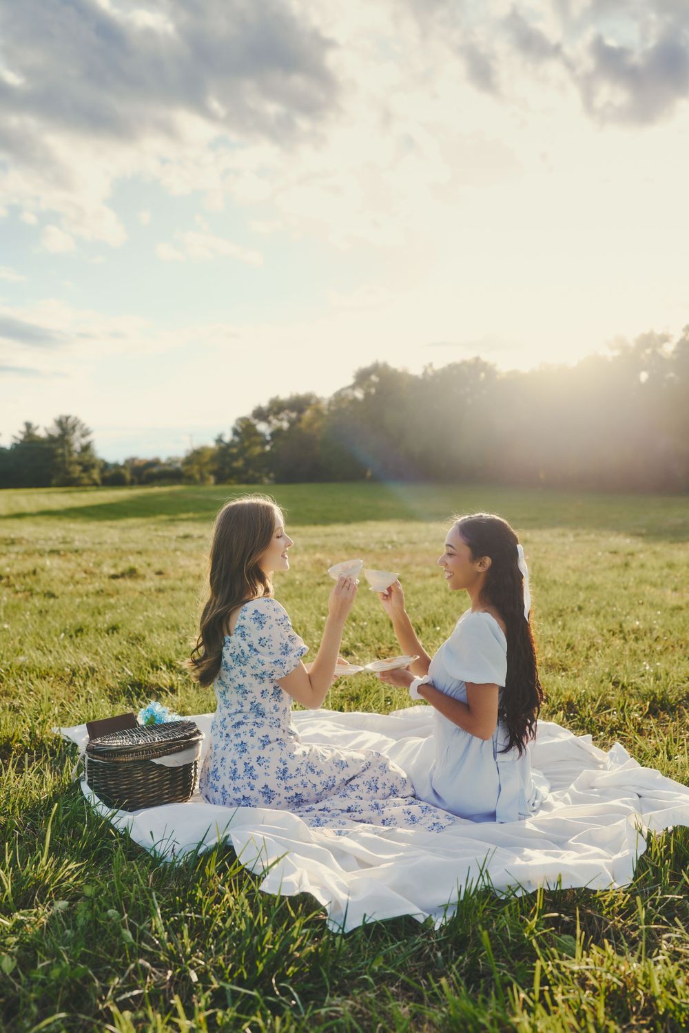 Two friends enjoying sunset picnic on blanket in grassy field with picnic basket.