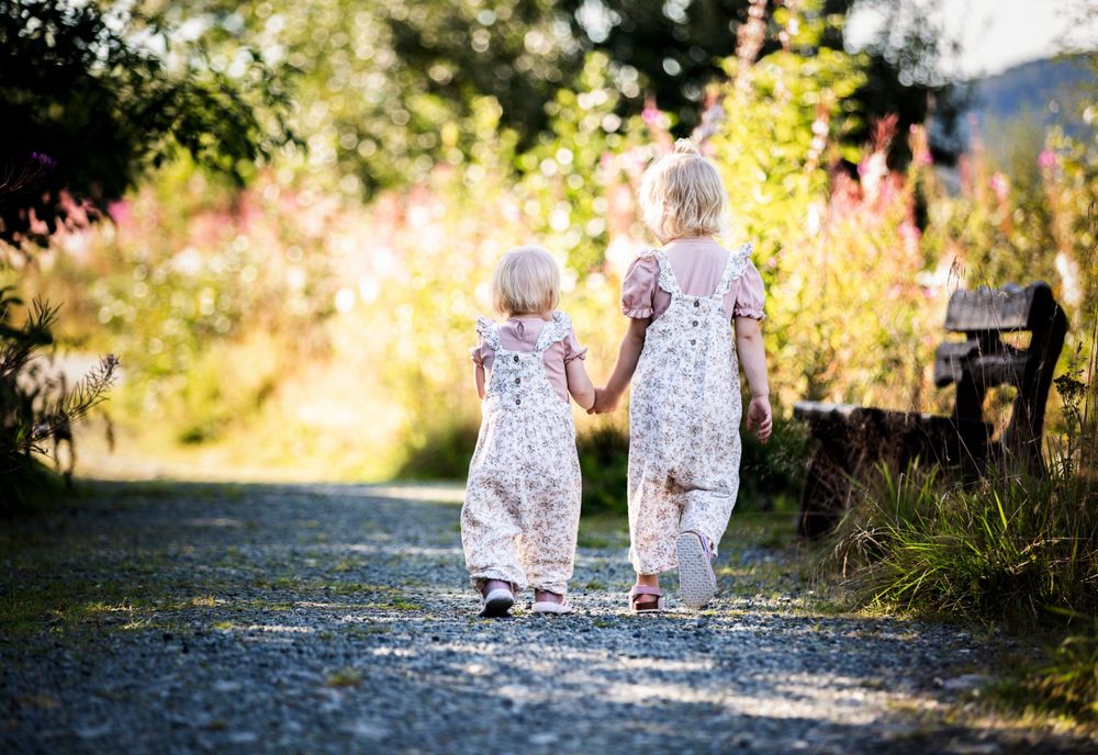 Two figures in matching dresses walk hand-in-hand down a sunlit path.
