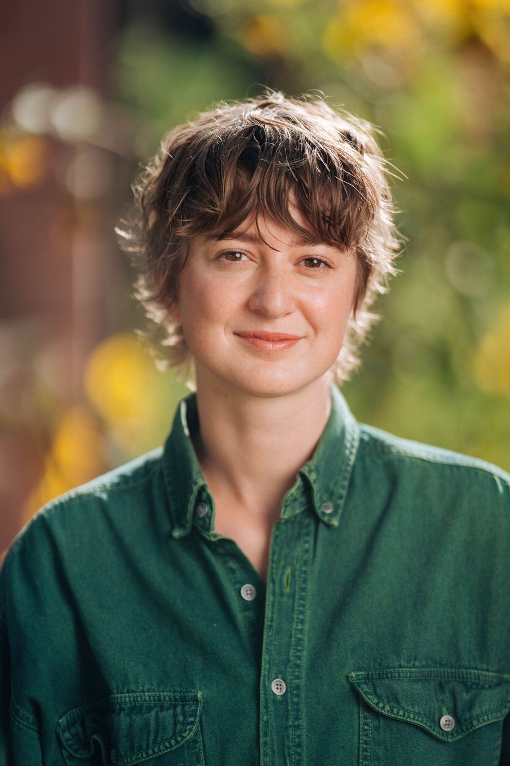 Young person with short brown hair smiling outdoors, wearing a green denim shirt with autumn foliage background.