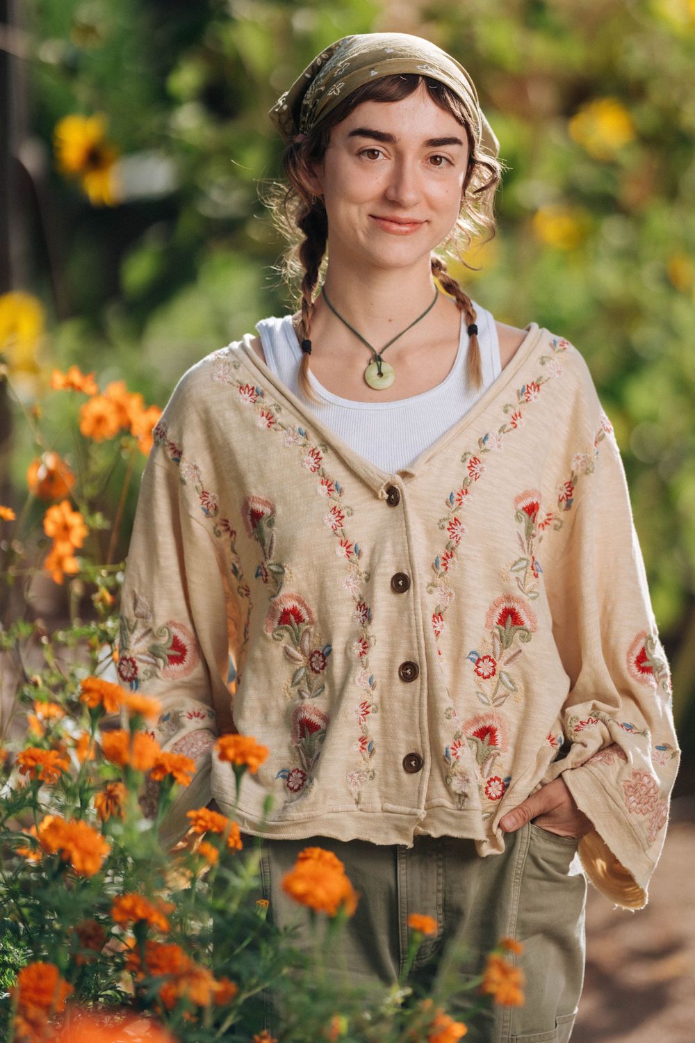 Young woman in floral embroidered cardigan smiling among orange marigolds in a sunlit garden.