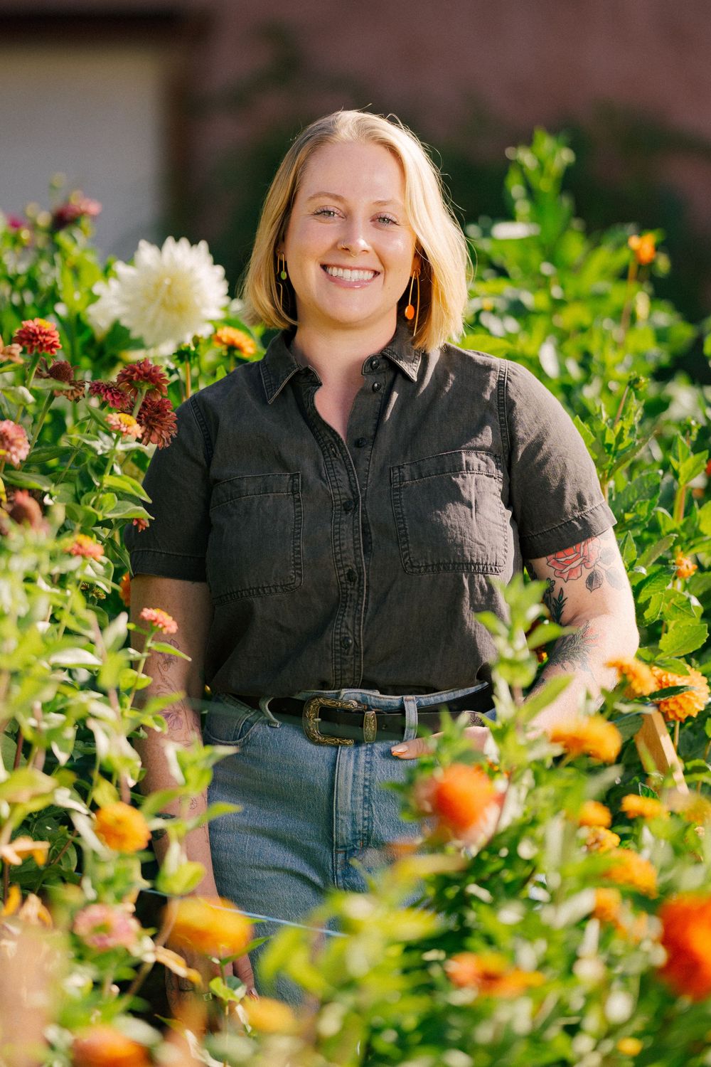 Smiling woman with tattoos standing among colorful garden flowers, wearing denim shirt and jeans outdoors.