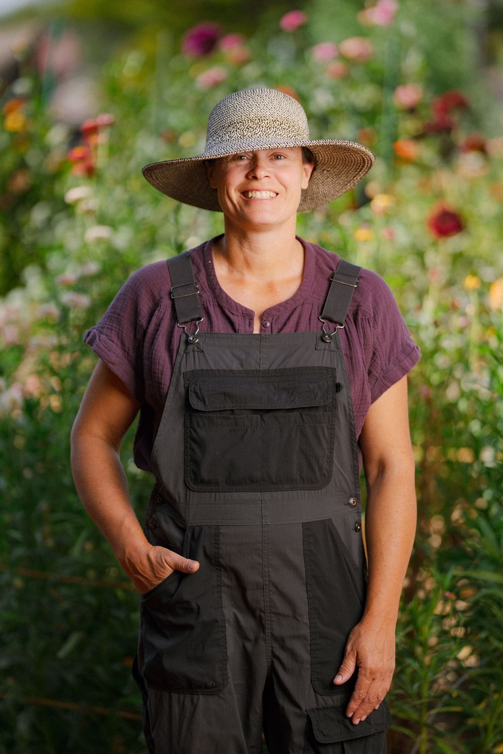 Smiling woman farmer in straw hat and overalls standing in a vibrant flower garden on a sunny day.