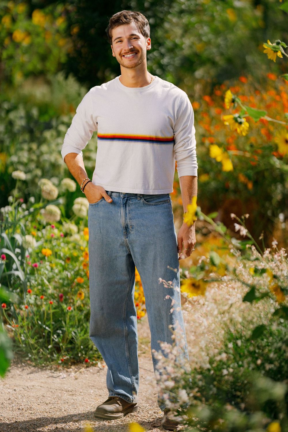 Smiling young man with mustache wearing white striped sweater and blue jeans standing in a colorful flower garden.