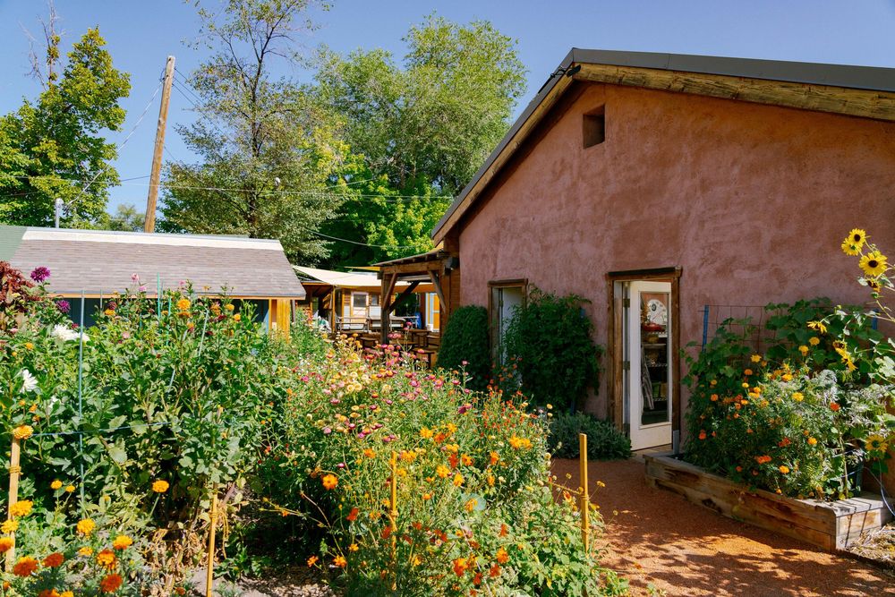Colorful garden with orange and yellow flowers beside a rustic adobe building surrounded by lush trees.