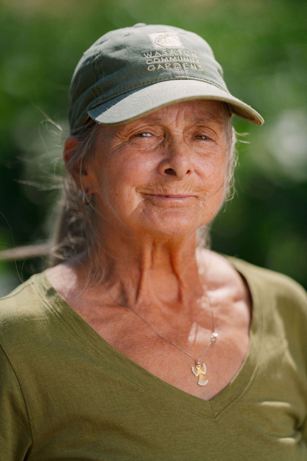 Elderly woman wearing a green community garden cap and olive t-shirt smiling outdoors in natural light.