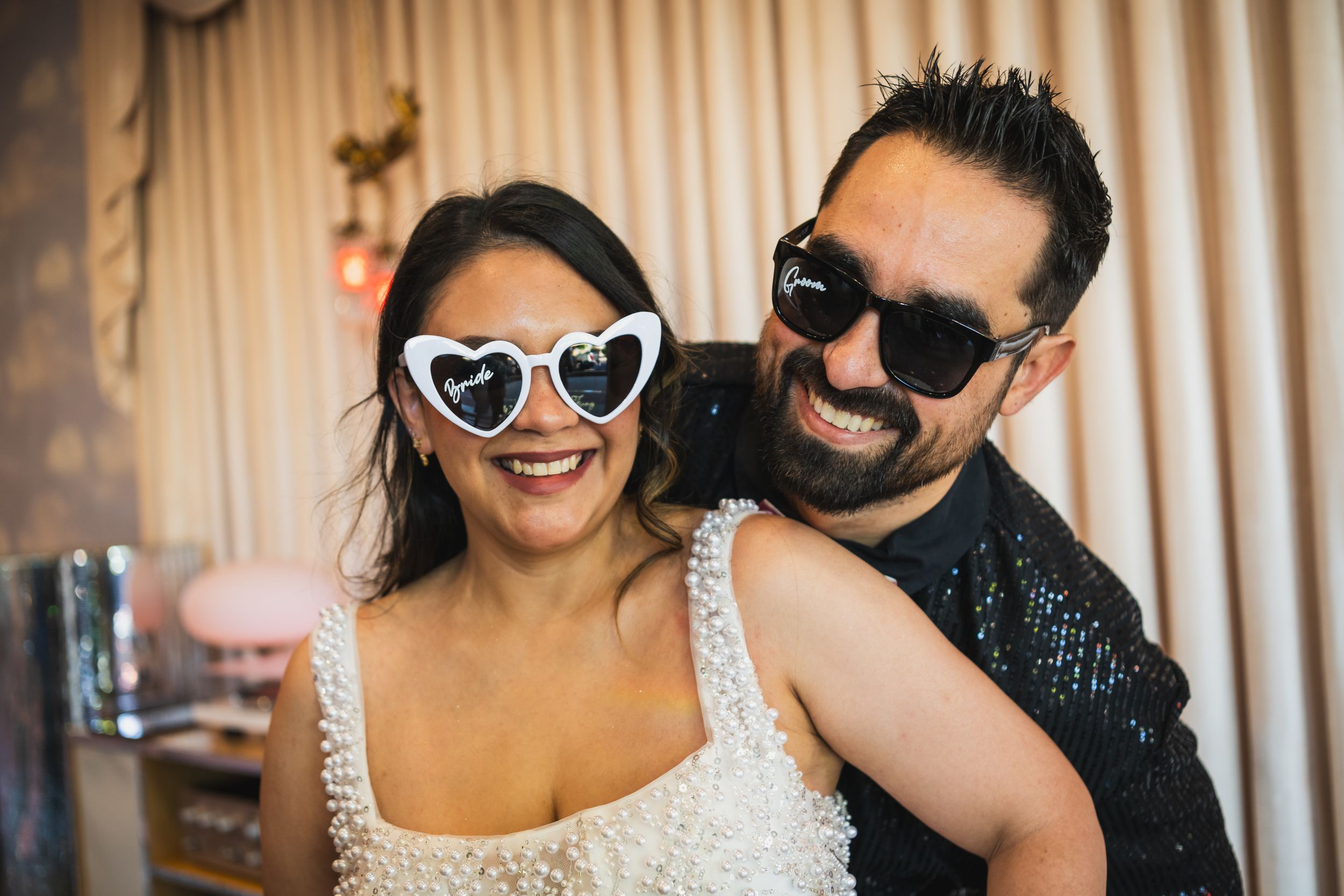 Happy couple wearing heart-shaped sunglasses smiling together at a festive indoor celebration.