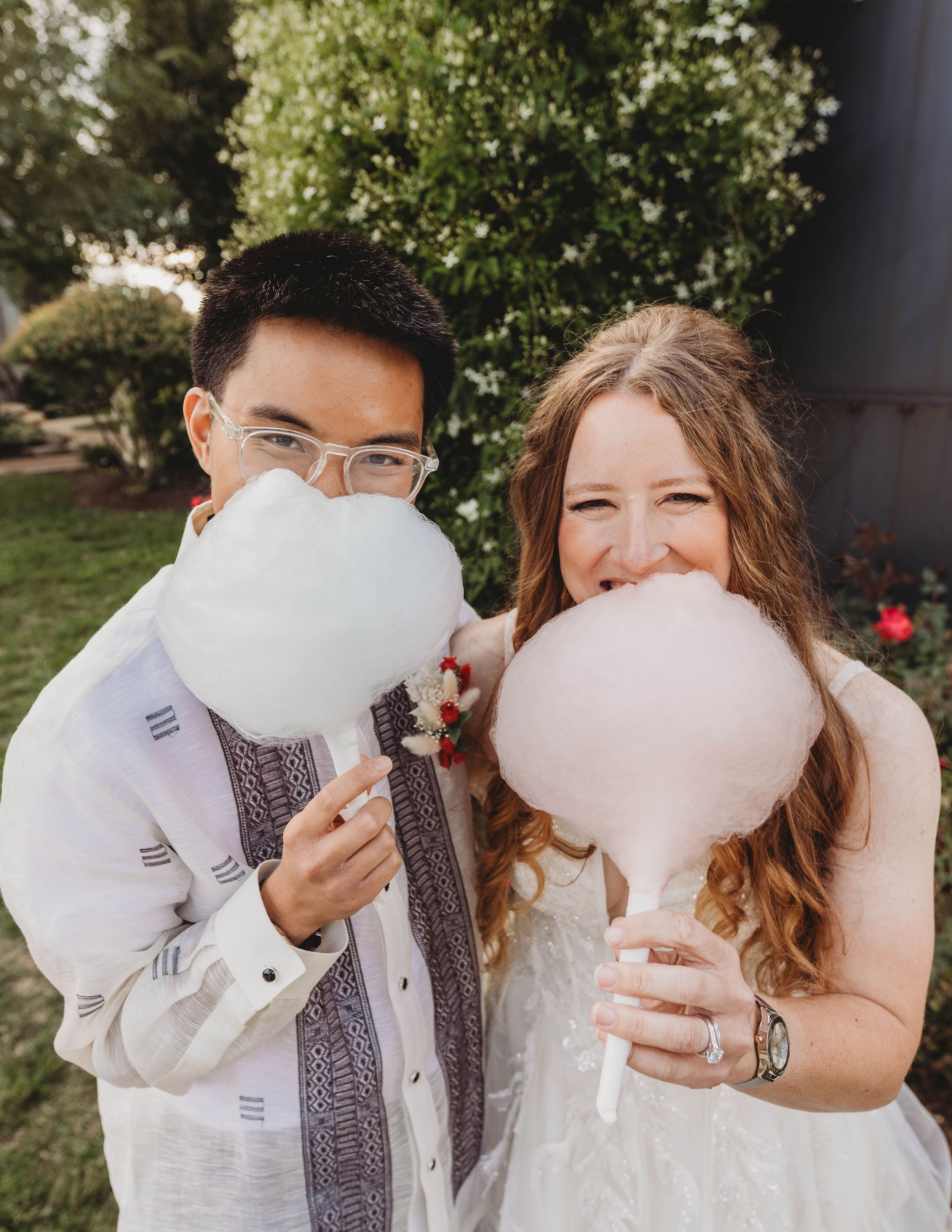 Playful couple eating cotton candy outdoors in wedding attire.