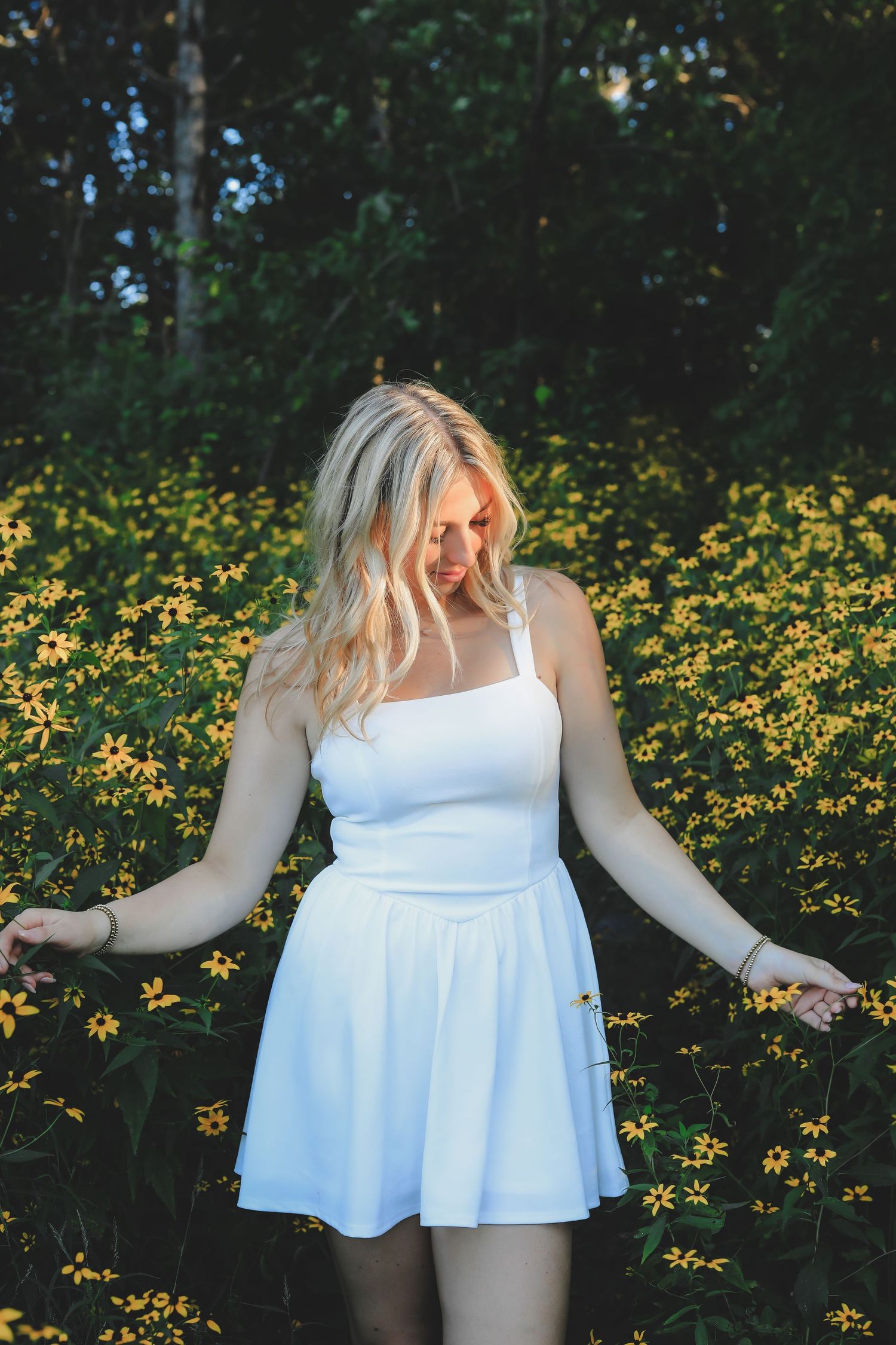 Blonde woman in white skater dress standing among bright yellow wildflowers in sunlit garden.