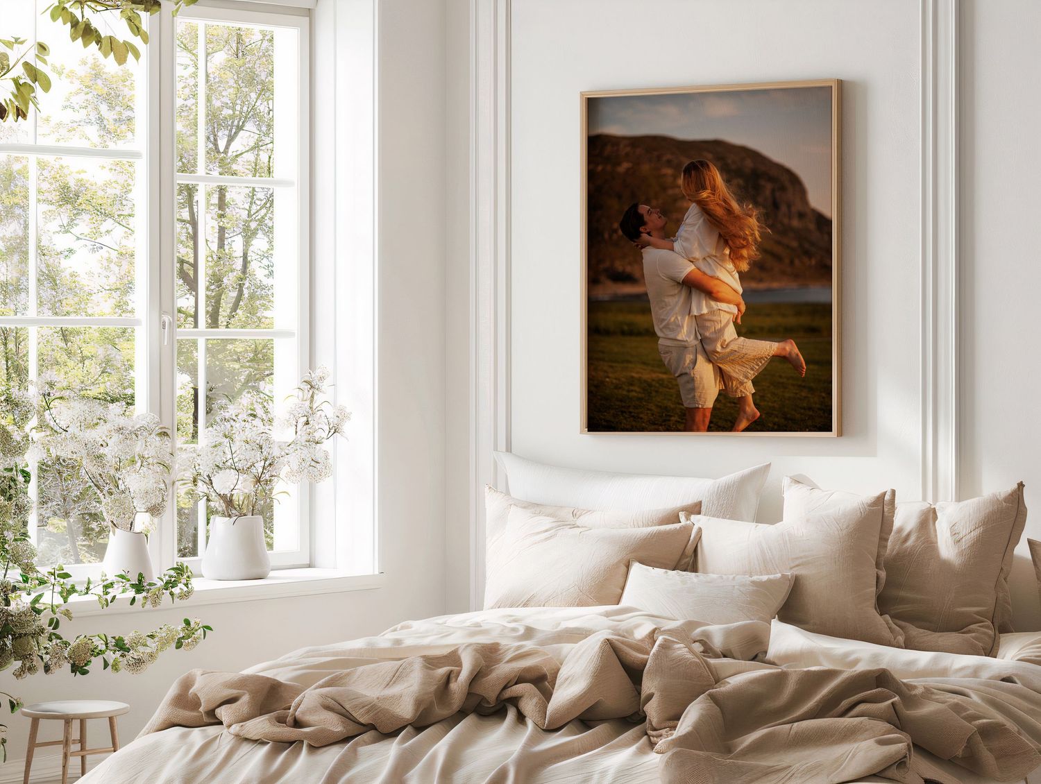 Cozy bedroom interior with beige bedding, bright window, and framed vintage-style artwork on white wall.