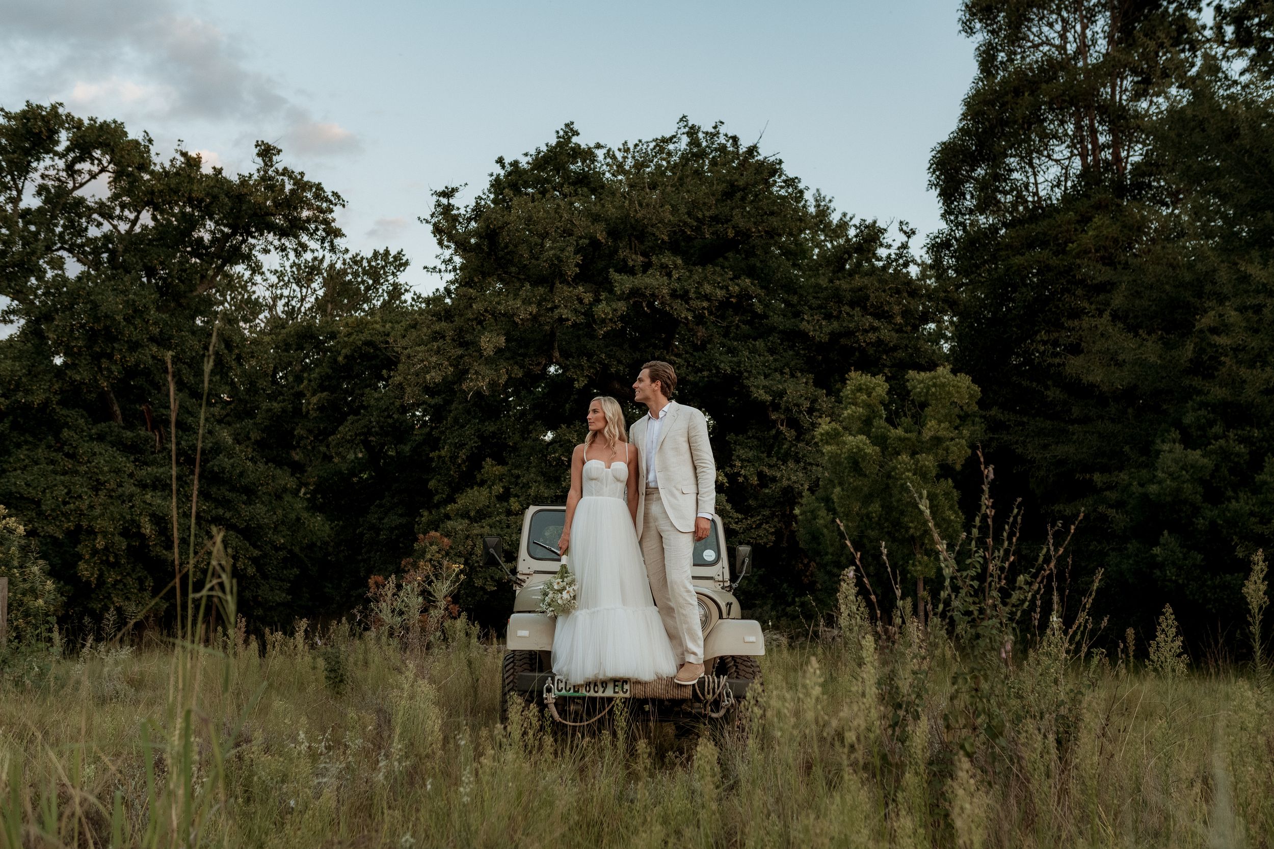 A couple in wedding attire walks through a serene green meadow with trees in the background at sunset.