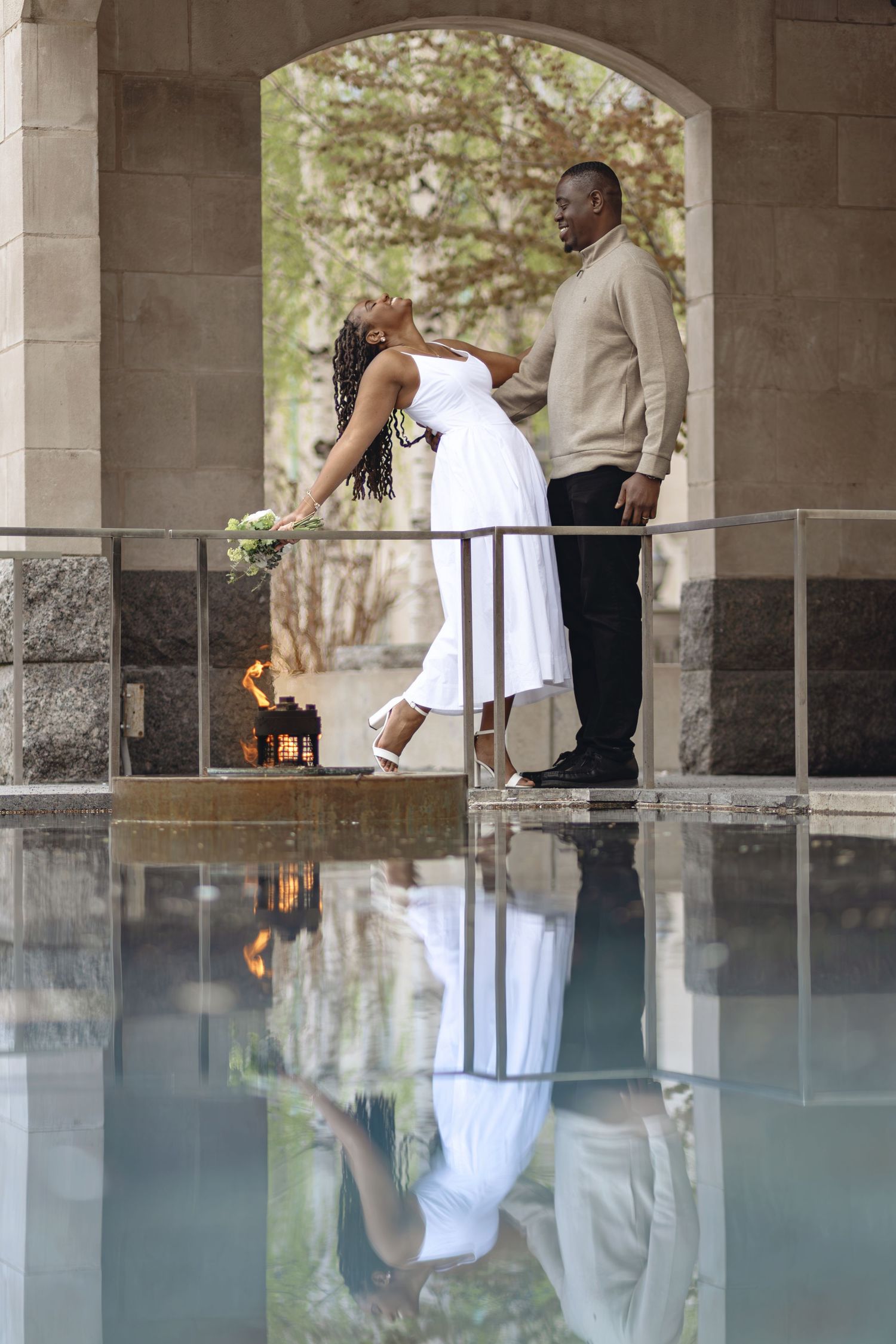 Bride in white dress and groom stand by reflective pool under stone arch with flaming lantern.