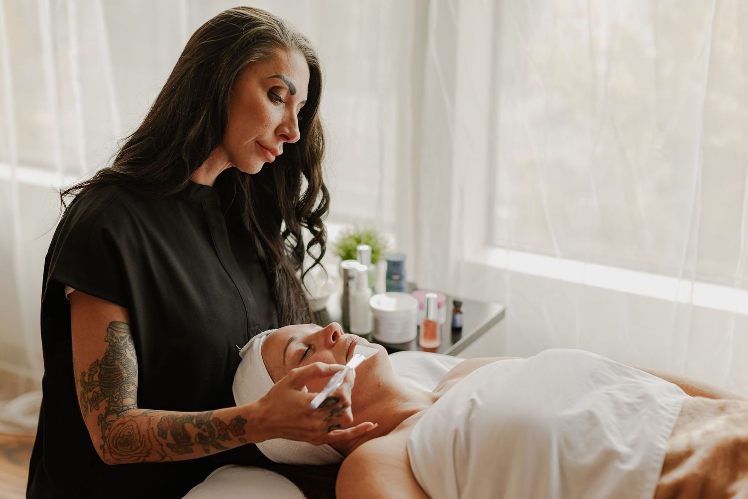 Esthetician with tattoos applying facial treatment to client in serene spa setting.