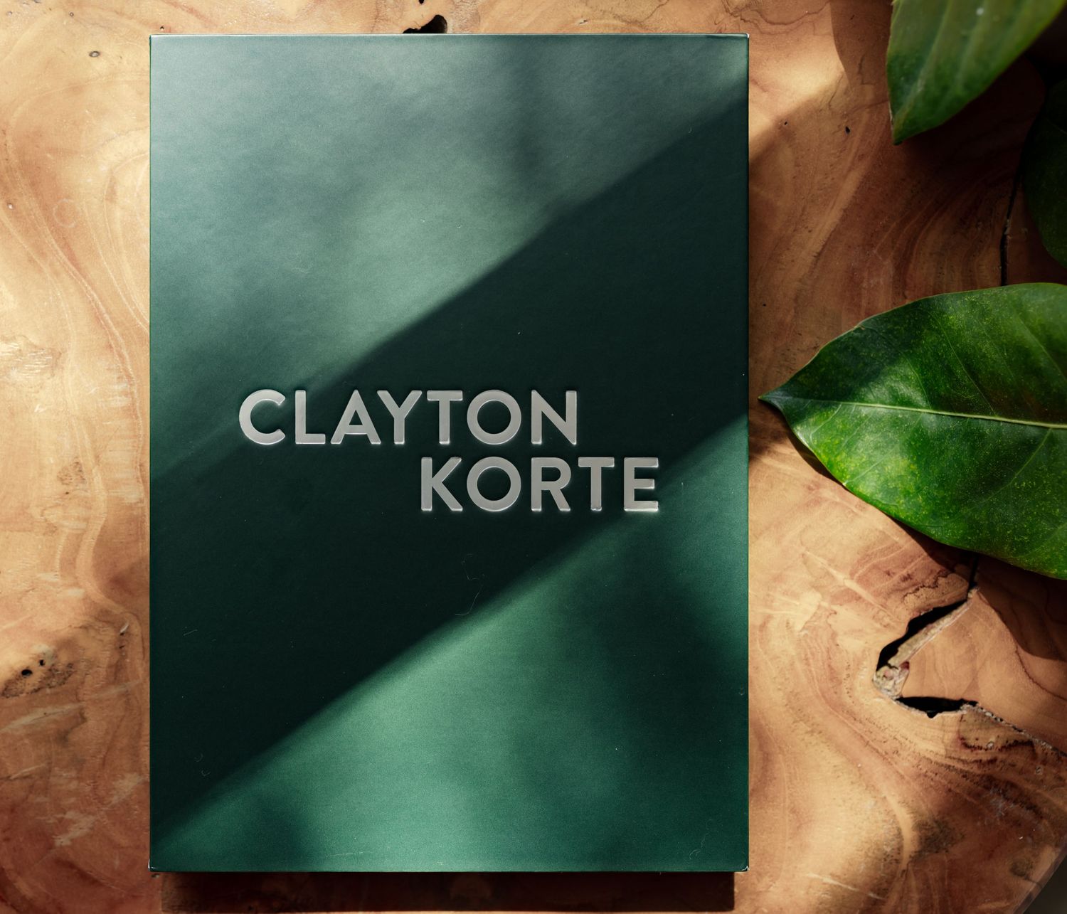 Dark green Clayton Korte branding card with silver text, placed on a wooden surface beside tropical leaves.