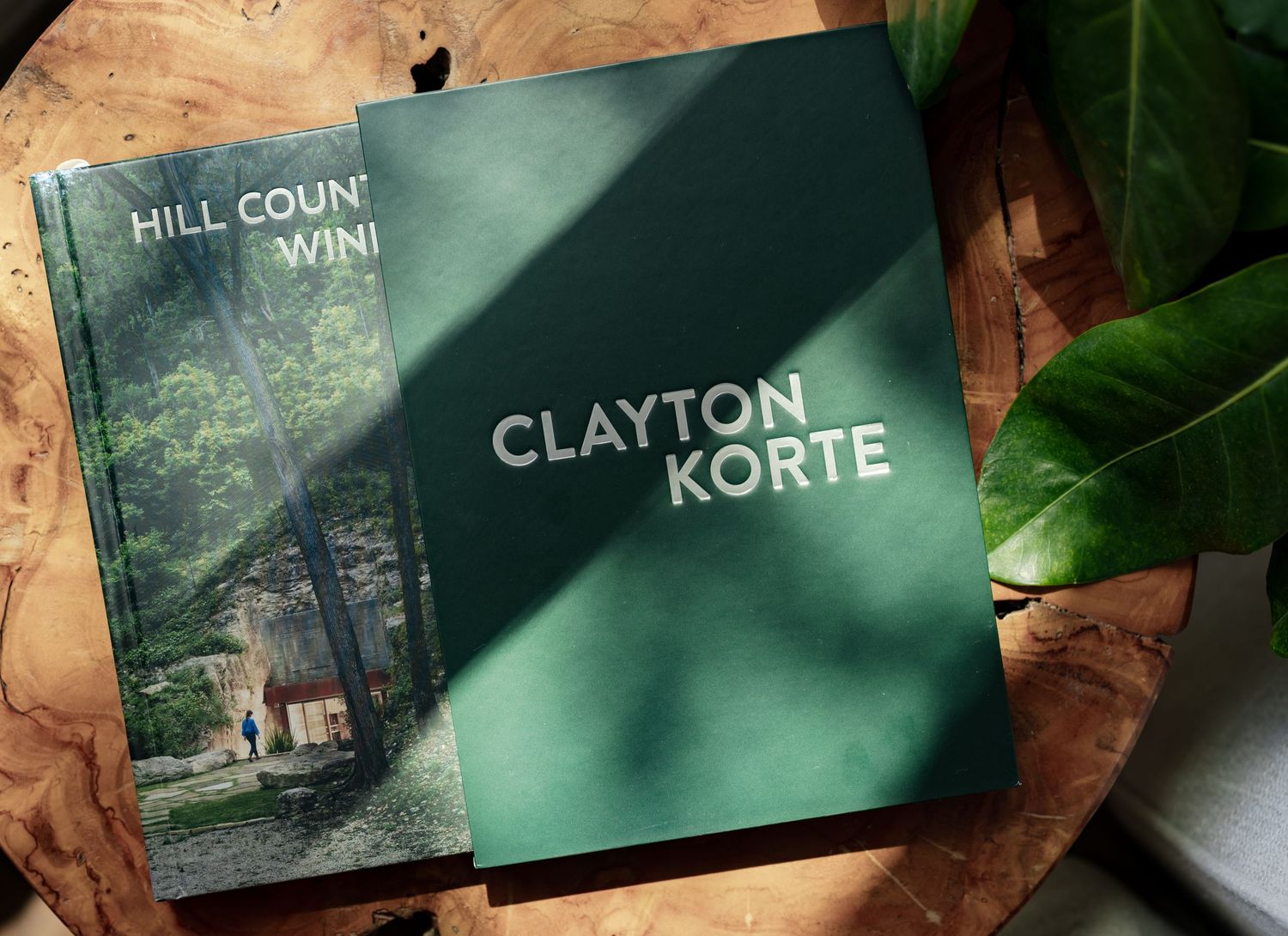Two books on a wooden table, including a green Clayton Korte architecture book and Hill Country Wine guide, with a plant nearby.