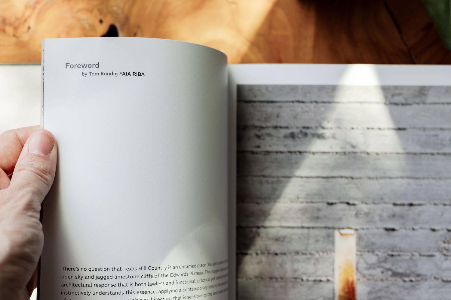 Hand holding open a book showing a foreword page beside a textured concrete wall with natural light.