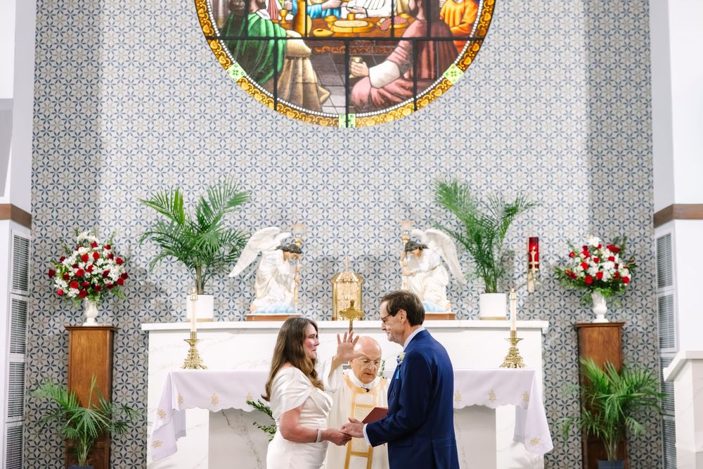 Couple exchanging vows at church altar with stained glass window, white flowers, and officiant during wedding ceremony.